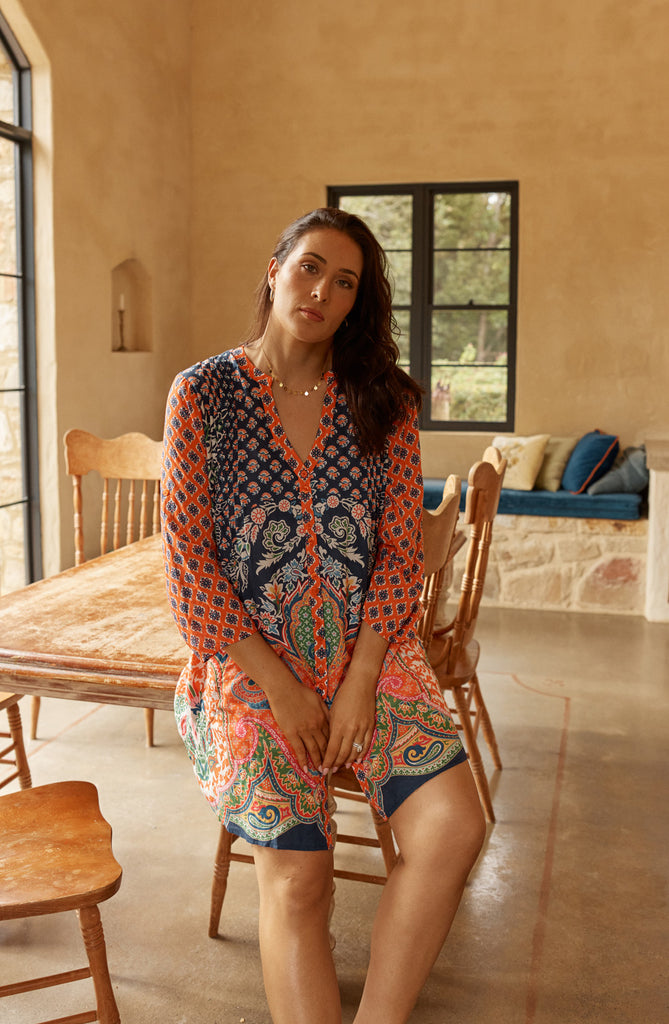 Model in the colourful patterned Lulalife Sarai button dress outfit sitting in a room with wooden furniture and a couch.